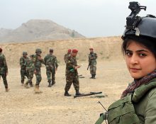 En bild på journalisten Najwa Alimi i kamouflage-kläder och en skyddshjälm som har en kamera fäst ovanpå. Hon tittar rakt in i kameran och i bakgrunden syns afghanska militärer.