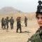 En bild på journalisten Najwa Alimi i kamouflage-kläder och en skyddshjälm som har en kamera fäst ovanpå. Hon tittar rakt in i kameran och i bakgrunden syns afghanska militärer.