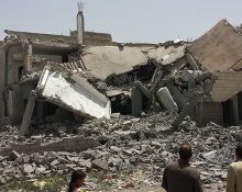 Ruinerna efter ett utbombat hus, med två vuxna och ett barn som står i förgrunden
