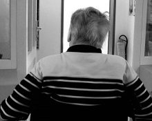 En äldre man i rullstol som sitter med ryggen mot kameran