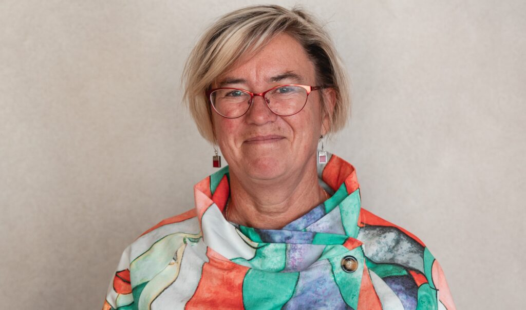 Porträtt av kvinna med glasögon, ljust hår i pagefrisyr och färgglad blus.