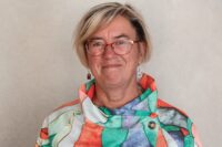 Porträtt av kvinna med glasögon, ljust hår i pagefrisyr och färgglad blus.